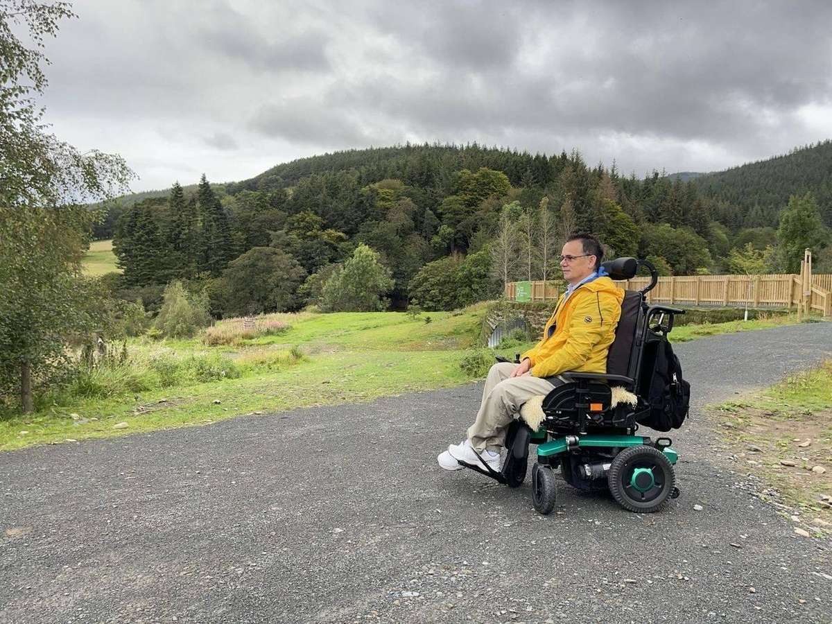 Paul sits in his bright green powerchair, looking out across a forest glade in Glentress Forest, Scottish Borders.
